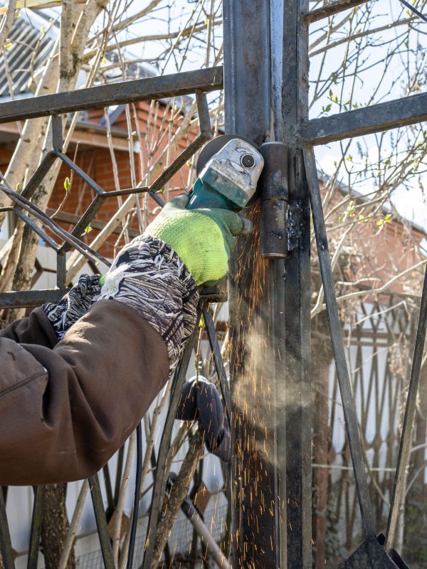 worker cutting hinge on gate of fence using electric grinder outdoors in village